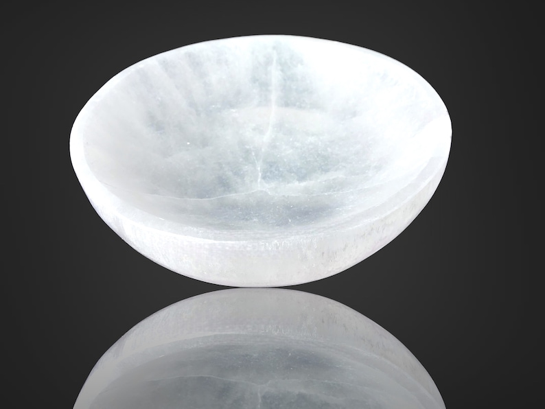 May include: A white selenite bowl with a smooth, polished surface. The bowl is slightly curved and has a reflective surface.