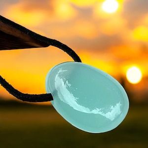 Può includere: Un ciondolo ovale azzurro realizzato in materiale traslucido, sospeso a un cordino nero. Il ciondolo è liscio e lucido, che riflette la calda luce del tramonto sullo sfondo. Il cordino sembra essere in pelle.