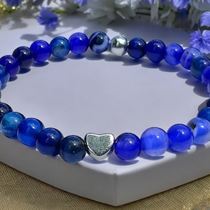 Könnte beinhalten: Ein blaues Perlenarmband mit einem silbernen Herzanhänger. Das Armband besteht aus runden, polierten Perlen in Blautönen. Der Herzanhänger befindet sich in der Mitte des Armbands. Das Armband wird auf einer weißen Oberfläche präsentiert.