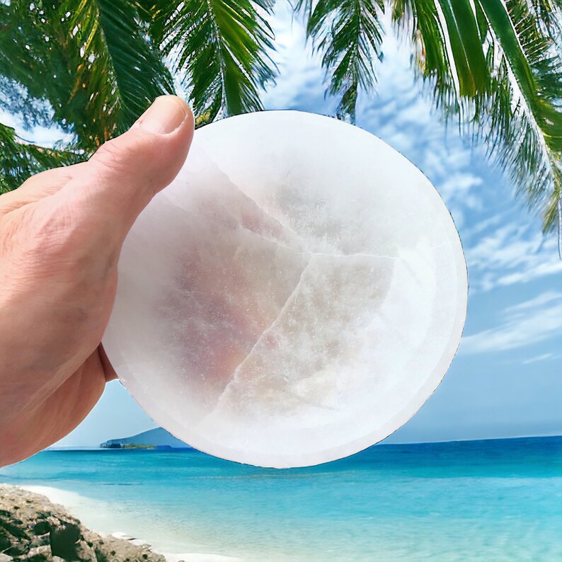 May include: A white selenite charging plate, a smooth, round disc with a slightly textured surface. The plate is held in a hand against a background of a tropical beach scene.