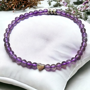Könnte beinhalten: Ein Amethyst-Perlenarmband mit einem silbernen Herzanhänger. Das Armband besteht aus kleinen, runden, lila Amethystperlen und einem silbernen Verschluss. Der Herzanhänger ist ein zentrales Merkmal des Armbands.