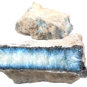 Könnte beinhalten: Zwei Stücke rohen, natürlichen blauen und weißen Agats. Das größere Stück ist hellbraun mit blauen Adern, die durch ihn verlaufen. Das kleinere Stück ist in zwei Hälften gebrochen und zeigt einen blauen und weißen Agat im Inneren.
