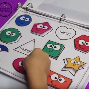 May include: A white binder with colorful shapes with faces on them. The shapes include a circle, a pentagon, a hexagon, a triangle, a square, a heart, a star, and a crescent moon. The words "triangle" and "heart" are written on the page.