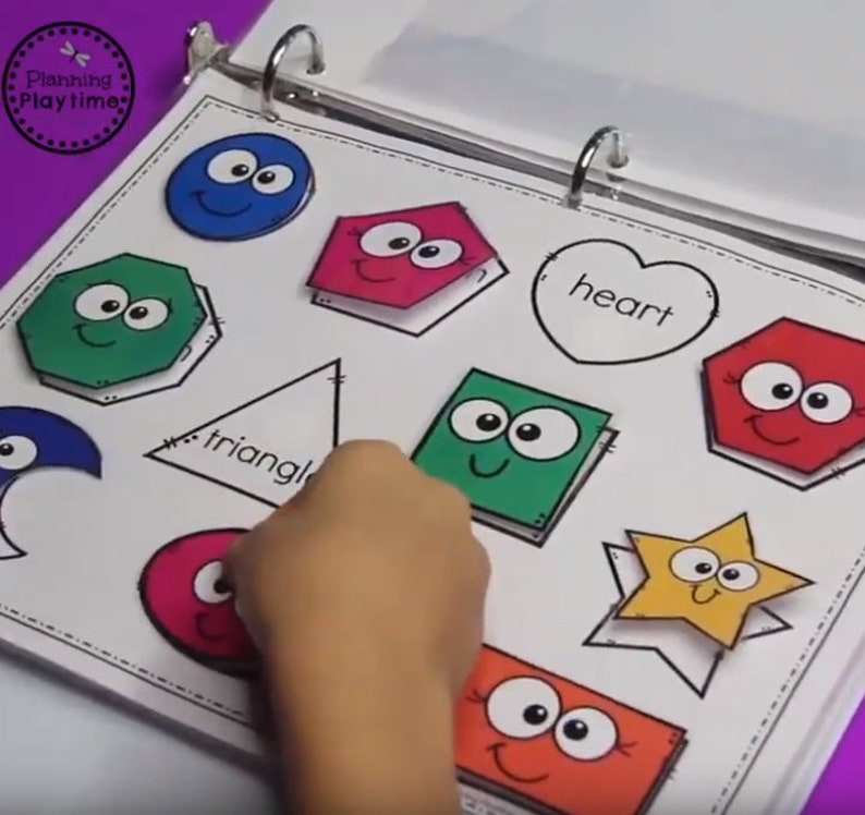 May include: A white binder with colorful shapes with faces on them. The shapes include a circle, a pentagon, a hexagon, a triangle, a square, a heart, a star, and a crescent moon. The words "triangle" and "heart" are written on the page.
