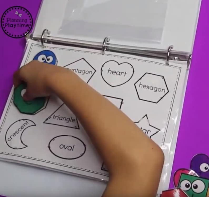 May include: A child's hand places a green pentagon shape onto a white sheet with black outlines of various shapes, including a heart, hexagon, triangle, crescent, oval, and star. The sheet is labeled with the names of each shape.