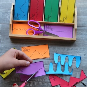 May include: A set of colourful paper strips with different cut-out lines for practising scissor skills. The strips are in a wooden tray with a pair of pink scissors. The strips are blue, red, green, yellow, and purple.