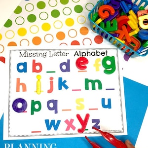 Puede incluir: Una tarjeta blanca con el texto "Missing Letter Alphabet" y las letras del alfabeto impresas en diferentes colores. Las letras a, b, d, e, f, g, h, i, j, k, m, o, p, q, s, u, w, x, y y z están impresas en la tarjeta. Hay espacios en blanco para las letras que faltan. Un par de pinzas rojas sostienen una letra o morada. La tarjeta está sobre una superficie azul con un fondo blanco con un patrón de círculos arcoíris.