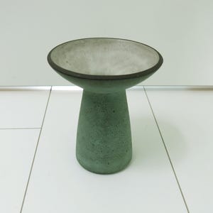 Puede incluir: Un cuenco con pedestal que presenta una base amplia y poco profunda y una base cilíndrica cónica. El exterior de la base y el borde tienen un acabado en verde salvia mate y texturizado, mientras que el interior del cuenco es de color gris claro.