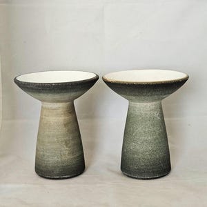May include: Two ceramic pedestal bowls with a flared top and a textured, gradient finish. The bowls transition from a dark base to a lighter, earthy tone, with a white interior. The design is minimalist and modern.