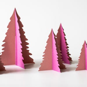 K&ouml;nnte beinhalten: Papier-Weihnachtsbaumdekorationen in verschiedenen Gr&ouml;&szlig;en. Die B&auml;ume sind braun und rosa, mit einem gefalteten Design. Die B&auml;ume sind auf einer wei&szlig;en Oberfl&auml;che angeordnet und schaffen eine festliche Weihnachtsdekoration.