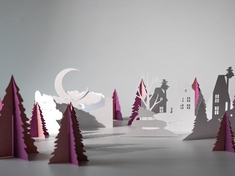 K&ouml;nnte beinhalten: Ein Papier-Diorama einer Winterlandschaft mit einem Halbmond, Wolken und silhouettierten H&auml;usern und B&auml;umen. Die B&auml;ume sind wei&szlig; und rosa, wodurch ein geschichteter Effekt entsteht. Die Szene ist vor einem hellgrauen Hintergrund.