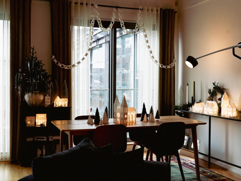 Peut inclure: Int&eacute;rieur chaleureux avec une table &agrave; manger en bois, des chaises et des maisons d&eacute;coratives en papier. Un petit sapin de No&euml;l et des guirlandes lumineuses ajoutent &agrave; l'ambiance festive. La pi&egrave;ce est &eacute;clair&eacute;e par un &eacute;clairage chaleureux.