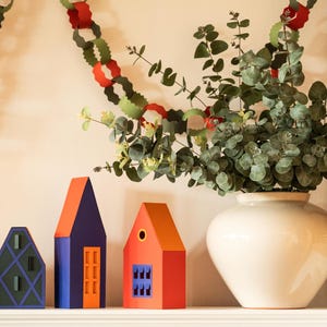 Puede incluir: Una composici&oacute;n decorativa en una estanter&iacute;a con casas de papel de colores, un jarr&oacute;n blanco con vegetaci&oacute;n y una guirnalda de papel. Las casas son naranjas, azules y rojas con dise&ntilde;os geom&eacute;tricos.