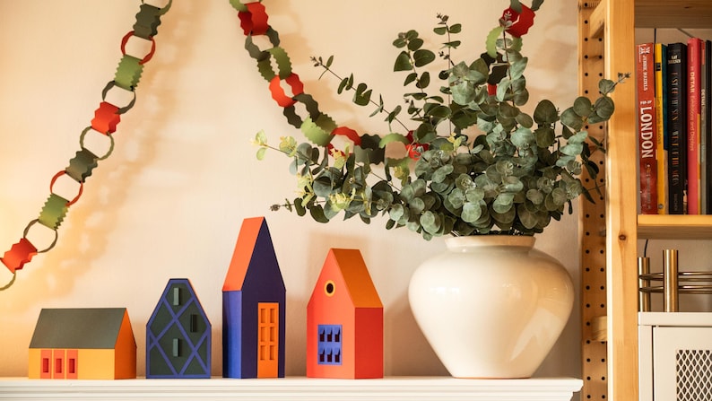 Puede incluir: Una composici&oacute;n decorativa en una estanter&iacute;a con casas de papel de colores, un jarr&oacute;n blanco con vegetaci&oacute;n y una guirnalda de papel. Las casas son naranjas, azules y rojas con dise&ntilde;os geom&eacute;tricos.