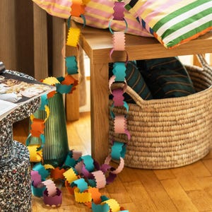 Puede incluir: Una colorida guirnalda de cadena de papel en tonos amarillo, naranja, rosa, morado y azul se extiende sobre una superficie de madera. Cojines a rayas y una cesta de mimbre en el fondo sugieren un tema festivo o decorativo.