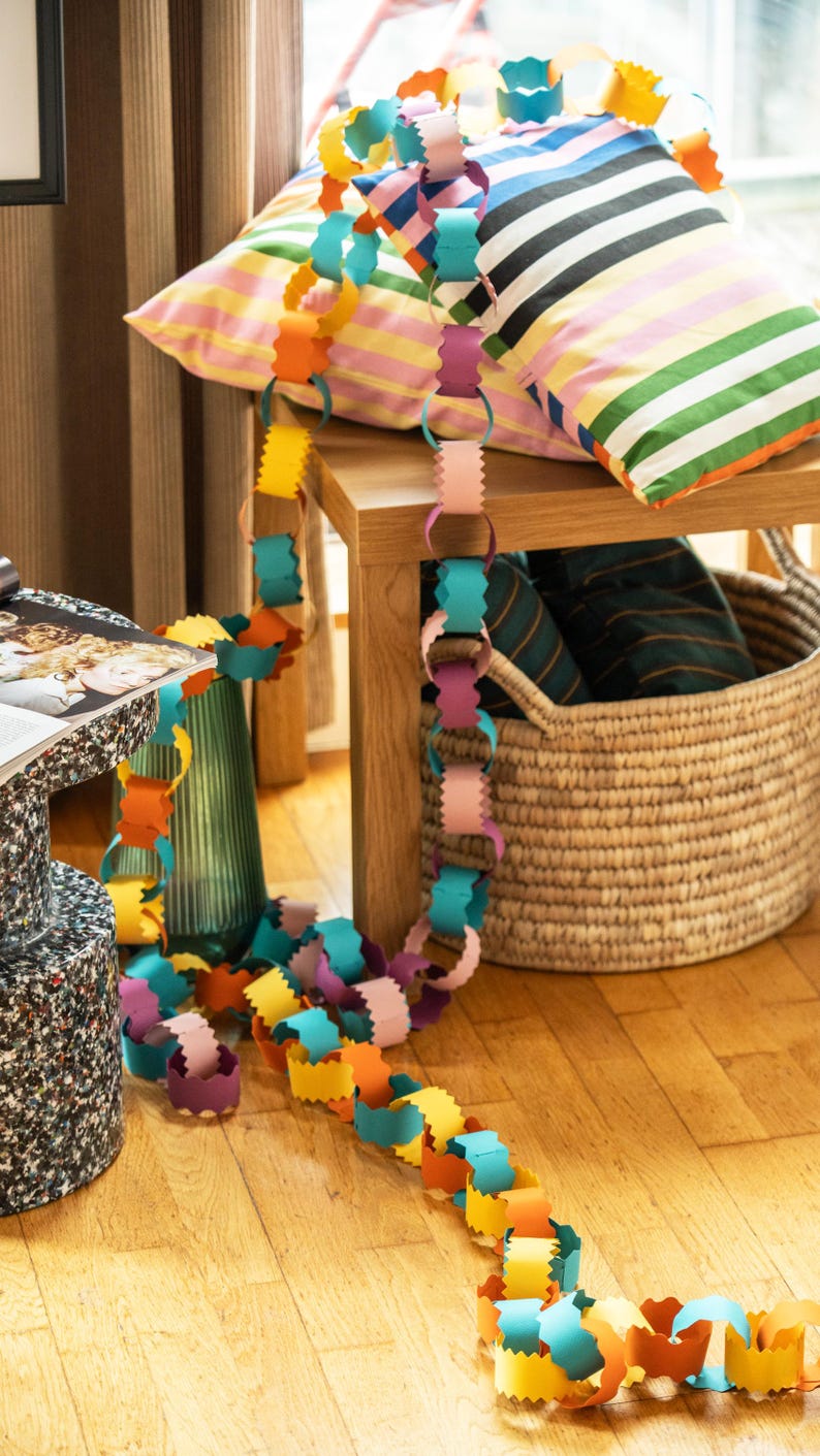 Puede incluir: Una colorida guirnalda de cadena de papel en tonos amarillo, naranja, rosa, morado y azul se extiende sobre una superficie de madera. Cojines a rayas y una cesta de mimbre en el fondo sugieren un tema festivo o decorativo.