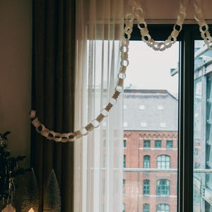 Peut inclure: Une fen&ecirc;tre avec des rideaux blancs transparents et des rideaux marron fonc&eacute;. Une guirlande de cha&icirc;ne en papier blanc est suspendue en haut. Une petite maison &eacute;clair&eacute;e et des arbres de No&euml;l sont pos&eacute;s sur une table.