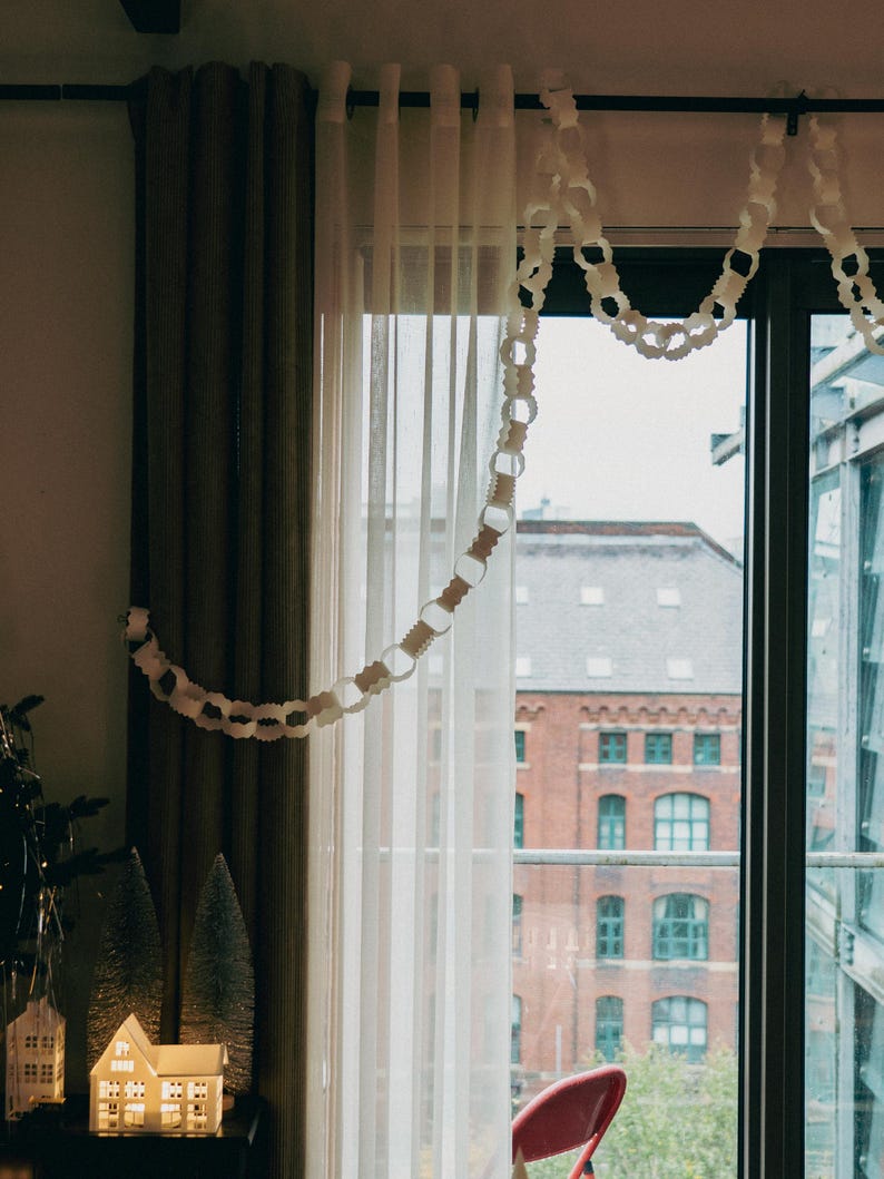 Peut inclure: Une fen&ecirc;tre avec des rideaux blancs transparents et des rideaux marron fonc&eacute;. Une guirlande de cha&icirc;ne en papier blanc est suspendue en haut. Une petite maison &eacute;clair&eacute;e et des arbres de No&euml;l sont pos&eacute;s sur une table.