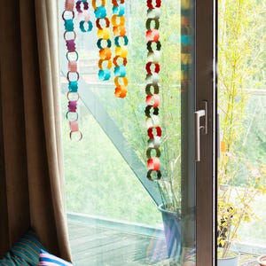 Puede incluir: Decoraciones de cadenas de papel de colores colgando frente a una ventana. Las cadenas son de varios colores, incluyendo turquesa, naranja y rojo. Cojines decorativos en una cesta est&aacute;n en primer plano. La ventana da a un balc&oacute;n con plantas.