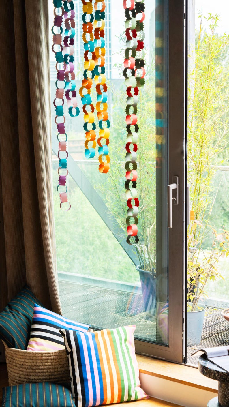 Puede incluir: Decoraciones de cadenas de papel de colores colgando frente a una ventana. Las cadenas son de varios colores, incluyendo turquesa, naranja y rojo. Cojines decorativos en una cesta est&aacute;n en primer plano. La ventana da a un balc&oacute;n con plantas.
