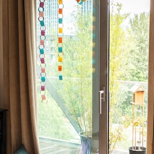 Puede incluir: Una colorida guirnalda de cadenas de papel cuelga frente a una ventana. La guirnalda presenta varios colores, incluyendo rosa, naranja, amarillo y azul. Cojines con dise&ntilde;os de rayas est&aacute;n en una cesta de mimbre cerca de la ventana.
