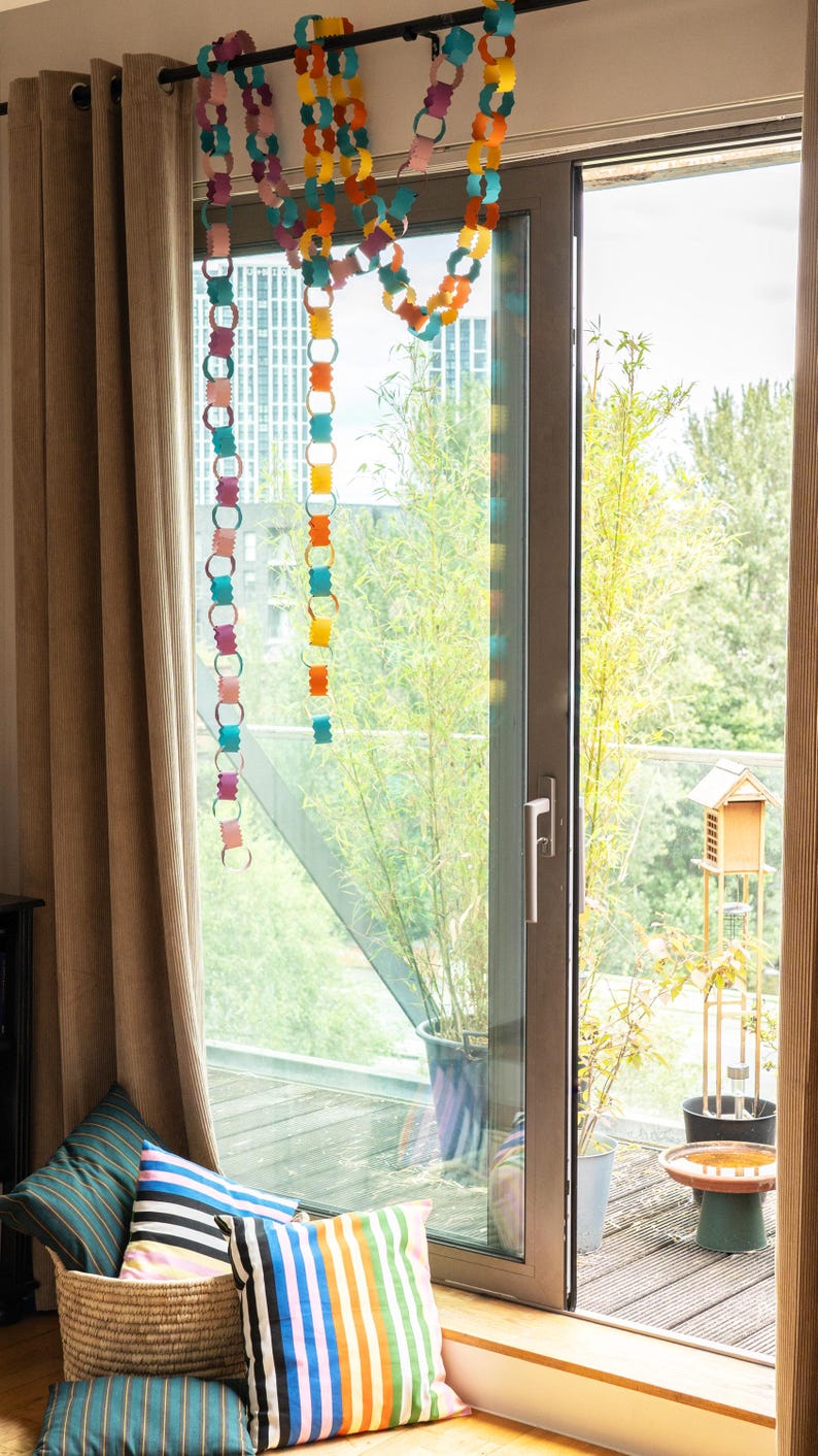 Puede incluir: Una colorida guirnalda de cadenas de papel cuelga frente a una ventana. La guirnalda presenta varios colores, incluyendo rosa, naranja, amarillo y azul. Cojines con dise&ntilde;os de rayas est&aacute;n en una cesta de mimbre cerca de la ventana.