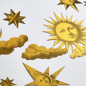 May include: Gold paper cut-outs of a crescent moon, sun, stars, and clouds. The moon, sun, and stars have faces drawn on them.