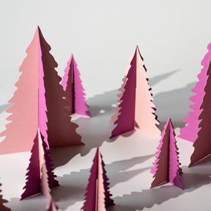 K&ouml;nnte beinhalten: Eine Ansammlung von rosa und magentafarbenen Weihnachtsb&auml;umen aus Papier, die auf einer wei&szlig;en Oberfl&auml;che angeordnet sind. Die B&auml;ume sind mit einer detaillierten, gezackten Kante geschnitten, wodurch eine wald&auml;hnliche Szene entsteht. Die Beleuchtung wirft Schatten und verst&auml;rkt den 3D-Effekt.