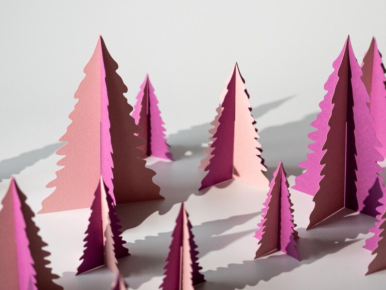 K&ouml;nnte beinhalten: Eine Ansammlung von rosa und magentafarbenen Weihnachtsb&auml;umen aus Papier, die auf einer wei&szlig;en Oberfl&auml;che angeordnet sind. Die B&auml;ume sind mit einer detaillierten, gezackten Kante geschnitten, wodurch eine wald&auml;hnliche Szene entsteht. Die Beleuchtung wirft Schatten und verst&auml;rkt den 3D-Effekt.