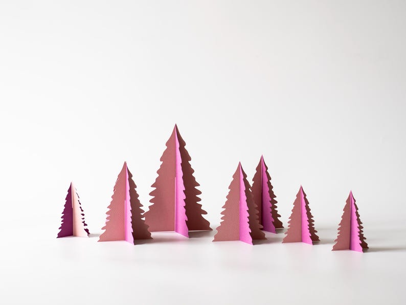 K&ouml;nnte beinhalten: Eine Sammlung rosa Weihnachtsb&auml;ume aus Papier in verschiedenen Gr&ouml;&szlig;en. Die B&auml;ume sind in einer Gruppe angeordnet, mit wei&szlig;em Hintergrund. Die B&auml;ume haben ein geschichtetes Design und erzeugen einen 3D-Effekt.