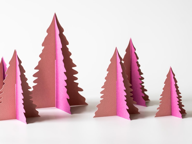 K&ouml;nnte beinhalten: Papier-Weihnachtsbaumdekorationen in verschiedenen Gr&ouml;&szlig;en. Die B&auml;ume sind braun und rosa, mit einem gefalteten Design. Die B&auml;ume sind auf einer wei&szlig;en Oberfl&auml;che angeordnet und schaffen eine festliche Weihnachtsdekoration.