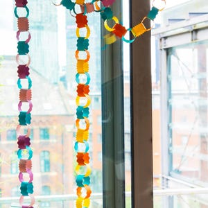 Puede incluir: Guirnalda de cadena de papel de colores colgando frente a una ventana. La guirnalda est&aacute; hecha de anillos de papel conectados en varios colores, incluyendo rosa, turquesa, naranja, amarillo y morado. La ventana da a un paisaje urbano.