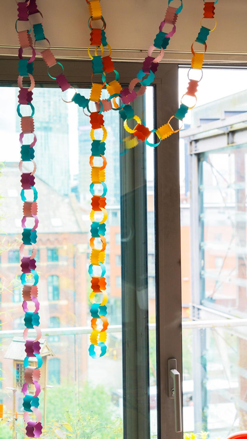 Puede incluir: Guirnalda de cadena de papel de colores colgando frente a una ventana. La guirnalda est&aacute; hecha de anillos de papel conectados en varios colores, incluyendo rosa, turquesa, naranja, amarillo y morado. La ventana da a un paisaje urbano.