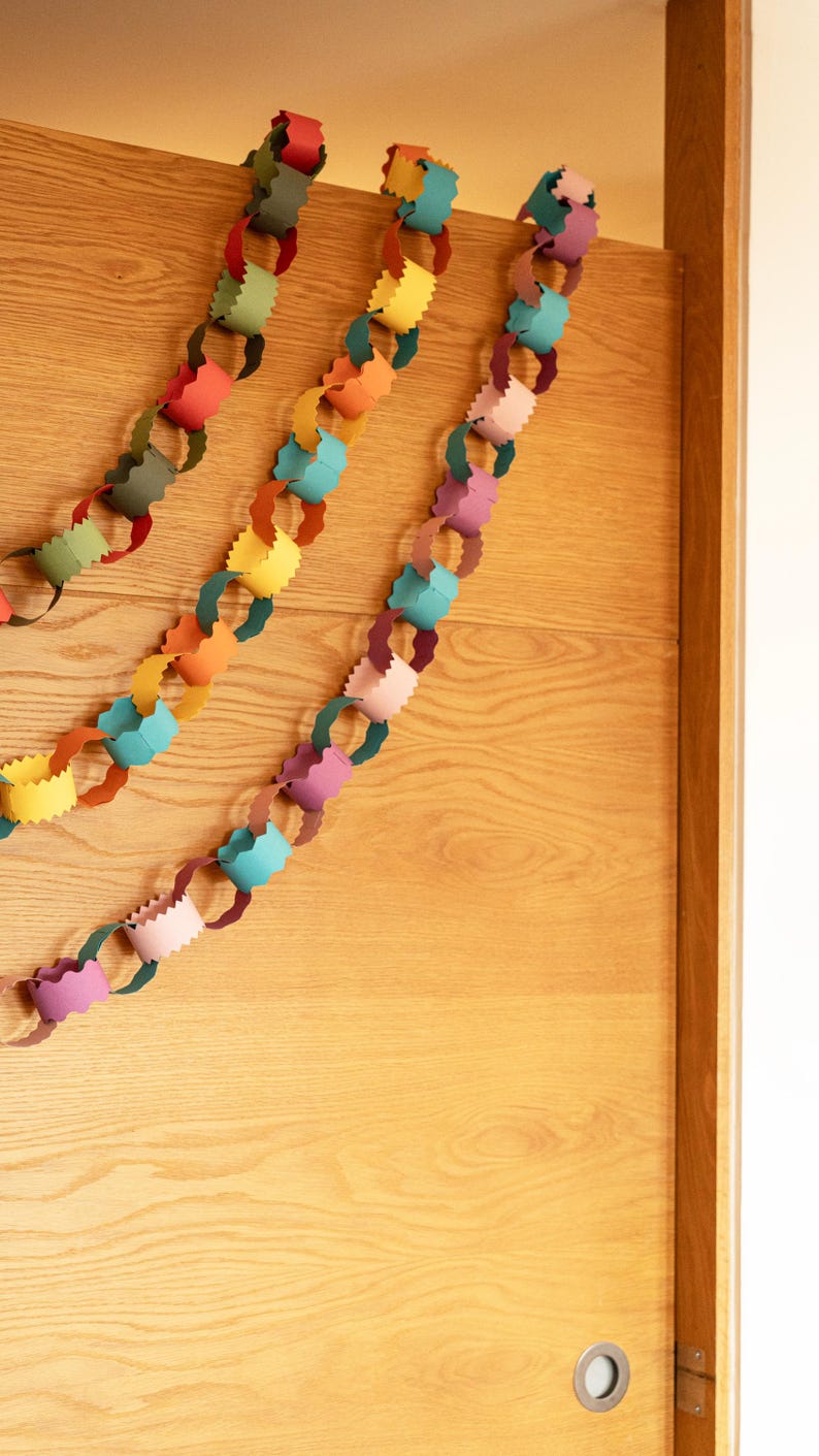 Puede incluir: Guirnalda de cadena de papel colorida en tonos rojo, verde, amarillo, naranja, azul y rosa, colgada en una puerta de madera clara. Los eslabones de papel est&aacute;n cortados con un borde decorativo, creando un aspecto festivo y l&uacute;dico. Ideal para decoraciones de fiesta.