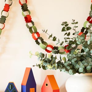 Puede incluir: Una exhibici&oacute;n festiva con una guirnalda de cadenas de papel en rojo, verde y blanco, colgando sobre casas de papel decorativas en azul, naranja y verde. Un jarr&oacute;n blanco contiene ramas de eucalipto.