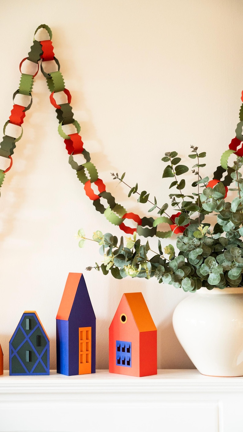 Puede incluir: Una exhibici&oacute;n festiva con una guirnalda de cadenas de papel en rojo, verde y blanco, colgando sobre casas de papel decorativas en azul, naranja y verde. Un jarr&oacute;n blanco contiene ramas de eucalipto.