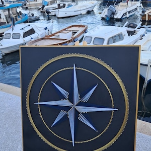 Puede incluir: Una pieza de arte de hilo con temática náutica, que representa una rosa de los vientos. La obra de arte utiliza hilo azul y blanco para la estrella de la brújula, e hilo dorado para los círculos, sobre un fondo negro. Se exhibe en un caballete de madera.