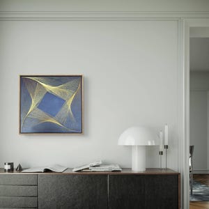 May include: A framed abstract art piece with a blue square center and gold string design. The artwork hangs on a white wall above a dark cabinet. A white mushroom-shaped lamp and a candle holder are on the cabinet.