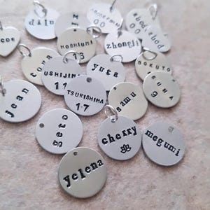 May include: A collection of small, silver-colored metal charms with various names and numbers stamped on them. Some charms are round, others are heart-shaped or rectangular. The charms have small loops for attaching to a chain or bracelet.