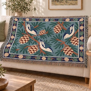 May include: A woven throw blanket featuring a repeating pattern of blue and white birds perched on pine branches with brown pinecones against a muted blue background, framed by a dark blue border with a pinecone and needle motif.