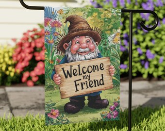 May include: A garden flag with a cartoon gnome holding a sign that says "Welcome my Friend". The gnome is wearing a brown hat and a blue shirt. The flag is surrounded by green grass and flowers.