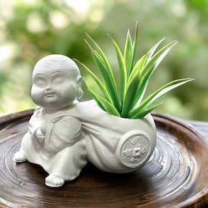 May include: A white matte planter shaped like a seated monk carrying a large sack on their back. The sack features a circular coin-like emblem on the side and contains a green succulent plant with pointed leaves, creating a functional decorative piece.