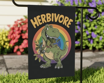May include: A black garden flag with a green dinosaur eating leaves. The dinosaur is in front of a rainbow and the text "HERBIVORE" is above the dinosaur.