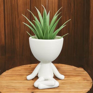 May include: White ceramic planter shaped like a person sitting in a meditative pose. The planter has a round, bowl-shaped top and holds a green succulent plant.