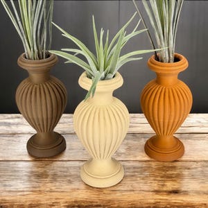 Pode incluir: Três vasos de cerâmica com designs nervurados em marrom, bege e laranja. Cada vaso está cheio de uma planta de ar verde.