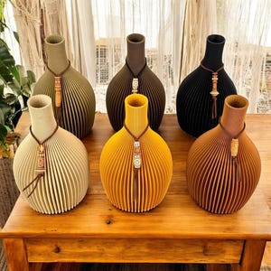 Op de afbeelding: Een verzameling van zes decoratieve vazen in verschillende kleuren, waaronder beige, olijfgroen, bruin, geel en zwart. Elke vaas heeft een uniek geribbeld ontwerp en is versierd met een decoratieve hanger en kwast. De vazen staan op een houten tafel.