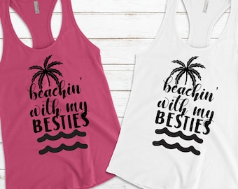 May include: Two tank tops with a palm tree and waves graphic and the text "beachin' with my besties". The tank top on the left is pink and the tank top on the right is white.