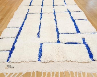 Puede incluir: Alfombra geométrica blanca y azul con borde de flecos. La alfombra tiene una textura esponjosa y está hecha de lana.