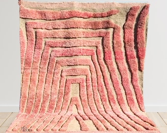May include: Pink and beige Moroccan rug with a geometric pattern of curved lines. The rug has a fringe border.