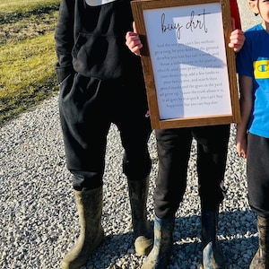 May include: A wooden sign with the text "buy dirt" and a poem about the importance of family, roots, and hard work. The sign is held by a child wearing black pants and blue rain boots.